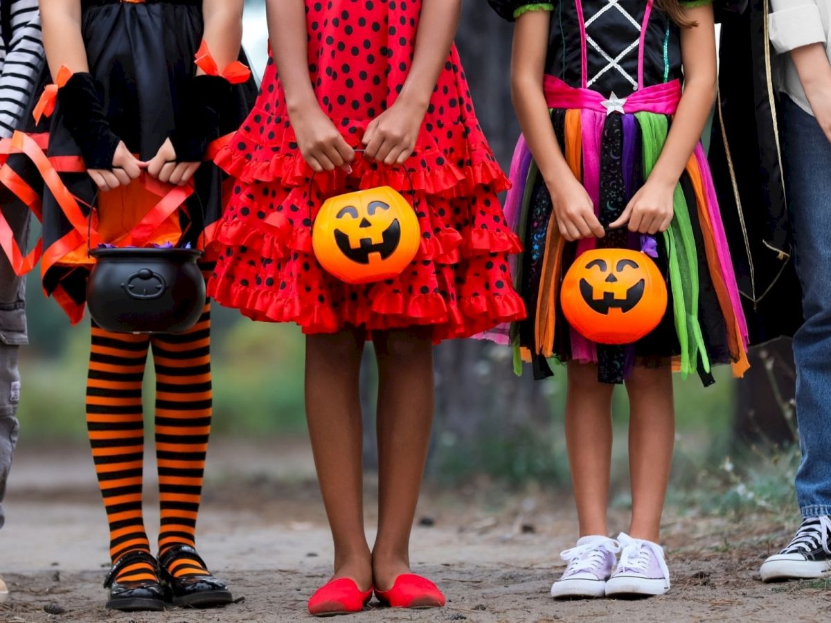 Kids in Halloween costumes hold orange jack‑o&rsquo;‑lantern candy pails outdoors.