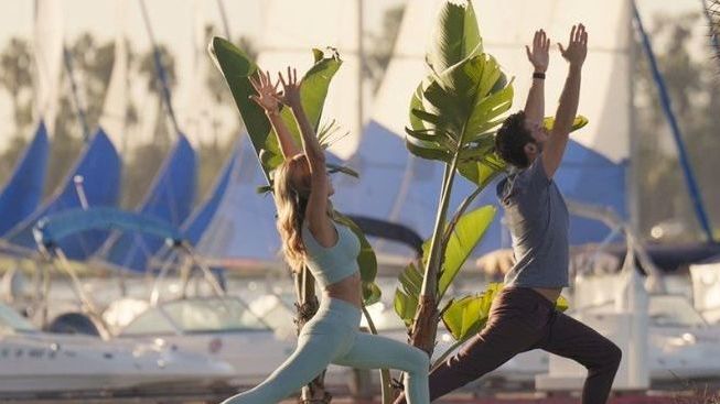 Two people stretch by palm trees in front of a marina with sailboats and tall flags on a sunny day.