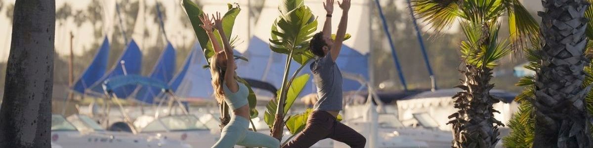 Two people stretch by palm trees in front of a marina with sailboats and tall flags on a sunny day.