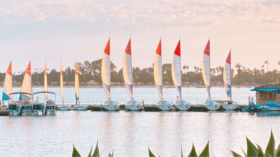 A row of sailboats with red-and-white sails is moored on a calm lake at sunset.