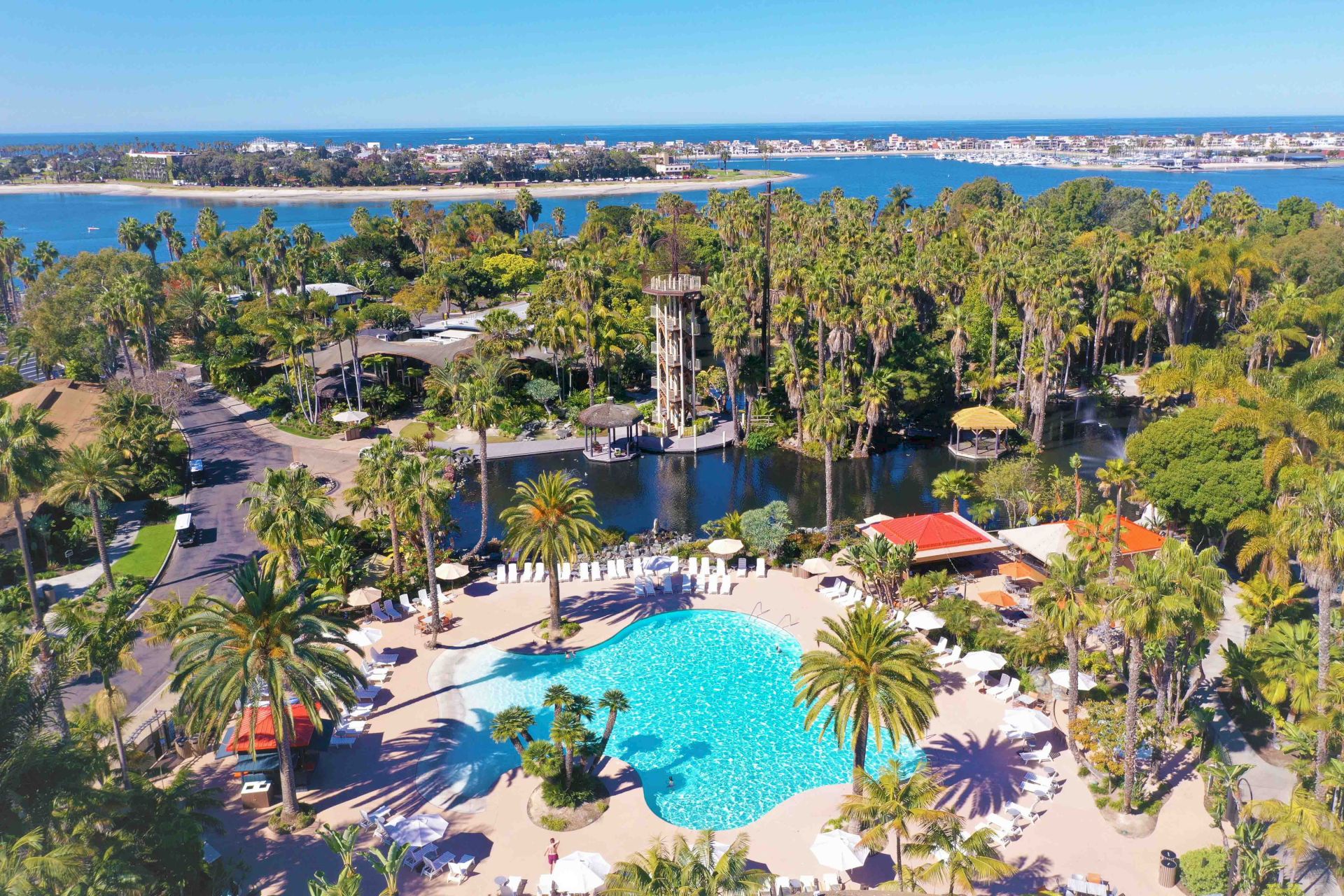 A sunny aerial view of a tropical beachfront resort with palm trees, a blue pool, and a marina overlooking the ocean.