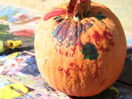 A round pumpkin painted with colorful faces sits on an art table with smeared paint around it.