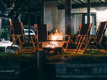 A backyard patio has wooden chairs around a glowing fire pit with flames burning under a roof.
