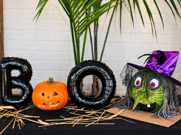 Halloween scene: foil balloons spell BOO, a carved jack-o&rsquo;-lantern sits by a grinning witch head, straw scattered everywhere.