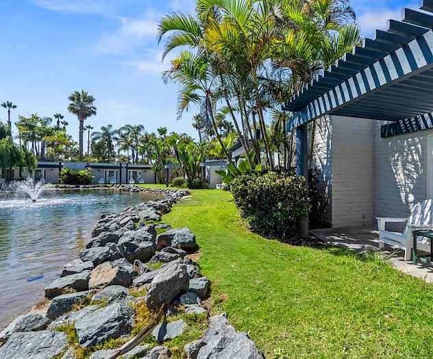 Tropical resort courtyard with a small pond, palm trees, and a white striped pergola beside a patio seating area.