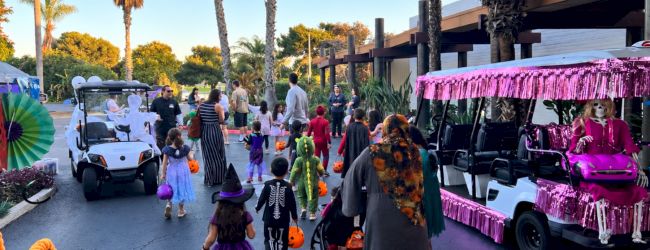 A Halloween parade with costumed kids walking between decorated carts under palm trees, with purple lights and candies.