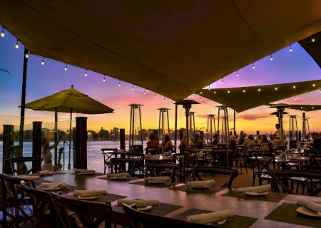 An outdoor waterfront dining patio at sunset with string lights, tables set for guests, umbrellas, and tall tiki heaters.
