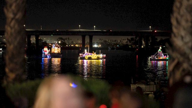 Night river parade: lit boats with colorful holiday figures cruise under a bridge, their reflections shimmering on dark water.