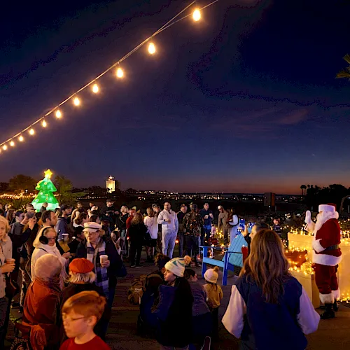 A festive crowd at a seaside promenade with string lights, a decorated tree, and people in holiday hats cheering.