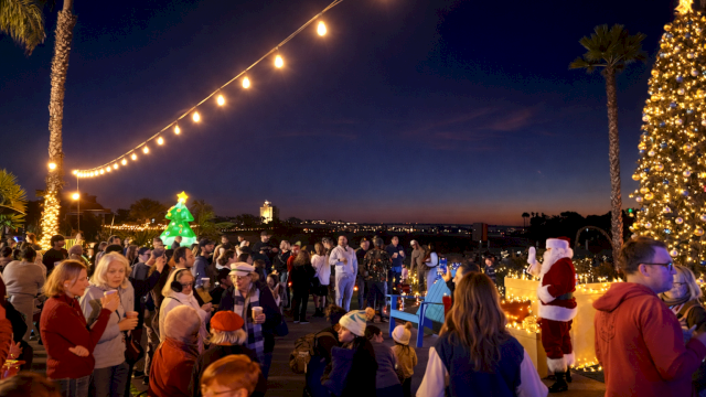 A festive crowd at a seaside promenade with string lights, a decorated tree, and people in holiday hats cheering.