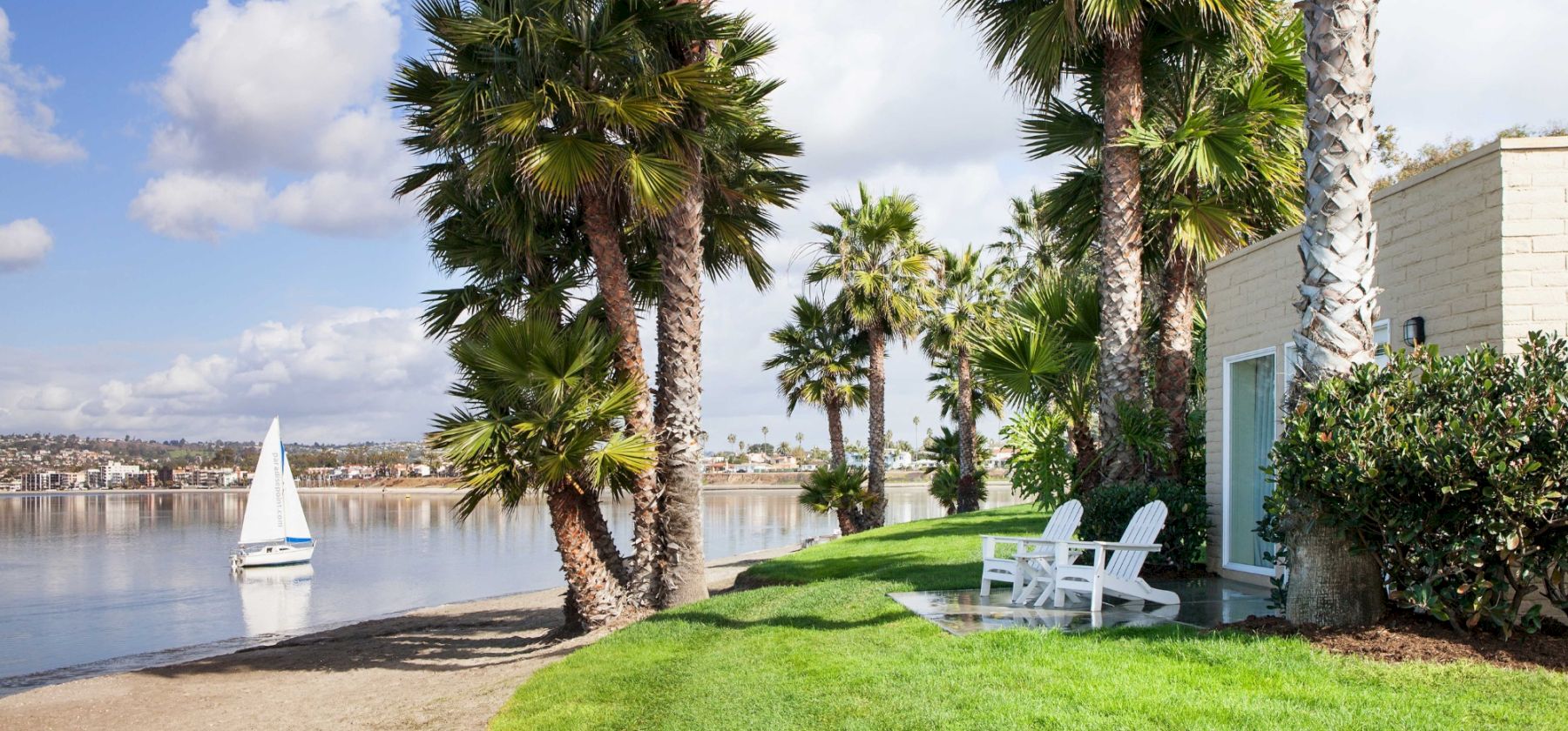 A sunny lakeside patio with tall palm trees, a green lawn, white Adirondack chairs, and a sailboat on calm water nearby.