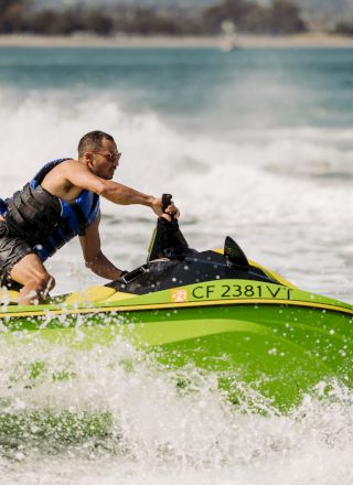 A man speeds on a green personal watercraft, wearing a life vest, blasting across choppy water with spray flying.