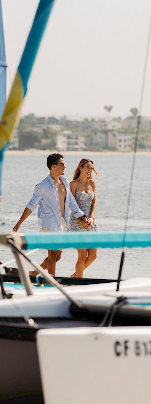 A couple walks and smiles on a sailboat deck near the water, framed by large blue sails with a coastal skyline in the distance.