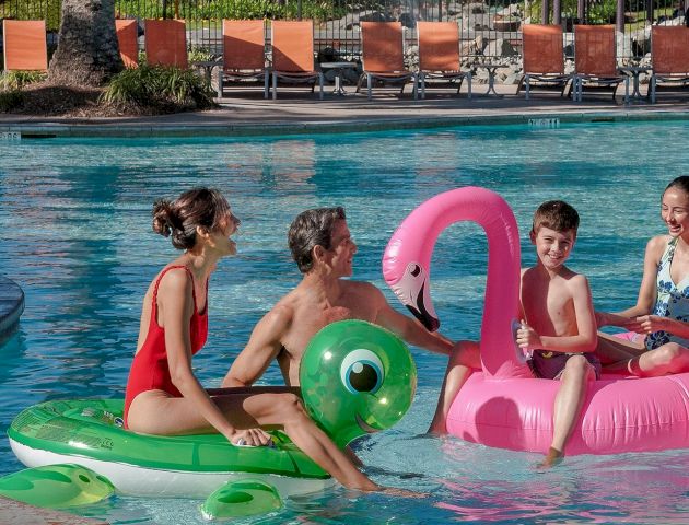 Kids playing in a sunny pool with colorful inflatable toys, including a green raft and pink flamingo float.