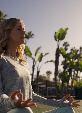 A woman practices yoga meditation outdoors in a sunny garden, palms swaying in the background.