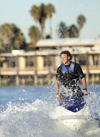 Two people ride jet skis, splashing water in a sunny seaside scene with a waterfront building and palm trees behind.