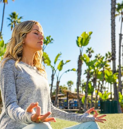 A woman sits cross‑legged in a tropical park, meditating on a mat among palm trees in sunny weather.