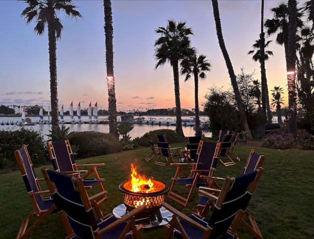 Dusk by a lake: palm trees, patio chairs around a glowing fire pit, and sailboats on calm water.