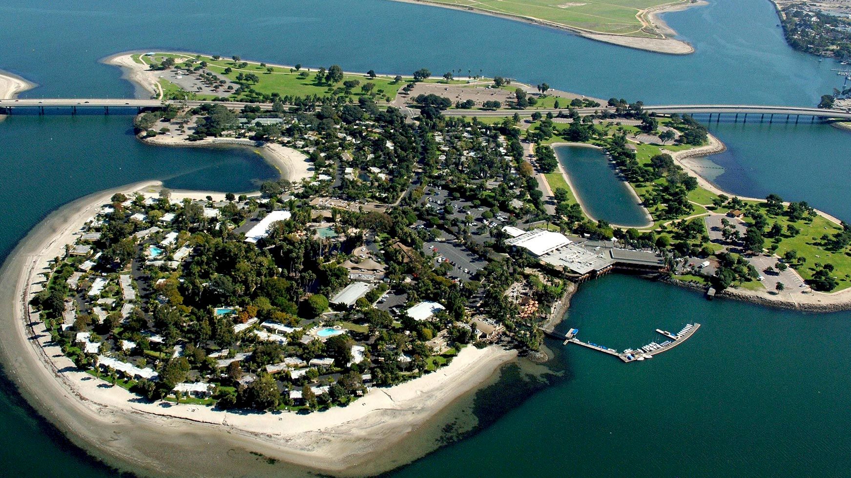 Aerial view of a riverside resort island with beaches, pools, and a marina, crossed by a bridge and waterways.