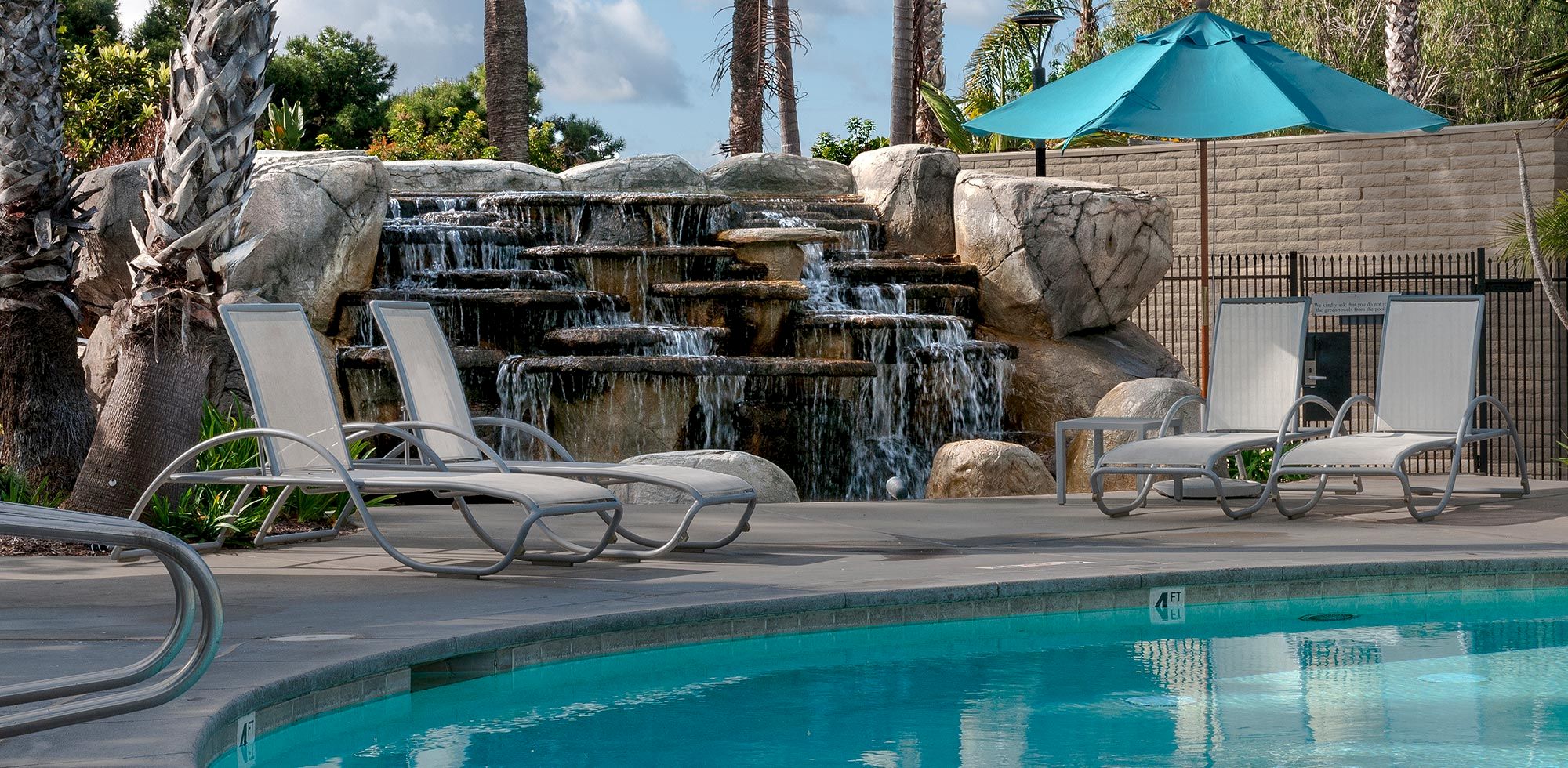 A sunny pool area with lounge chairs and a cascading rock waterfall under a teal umbrella.