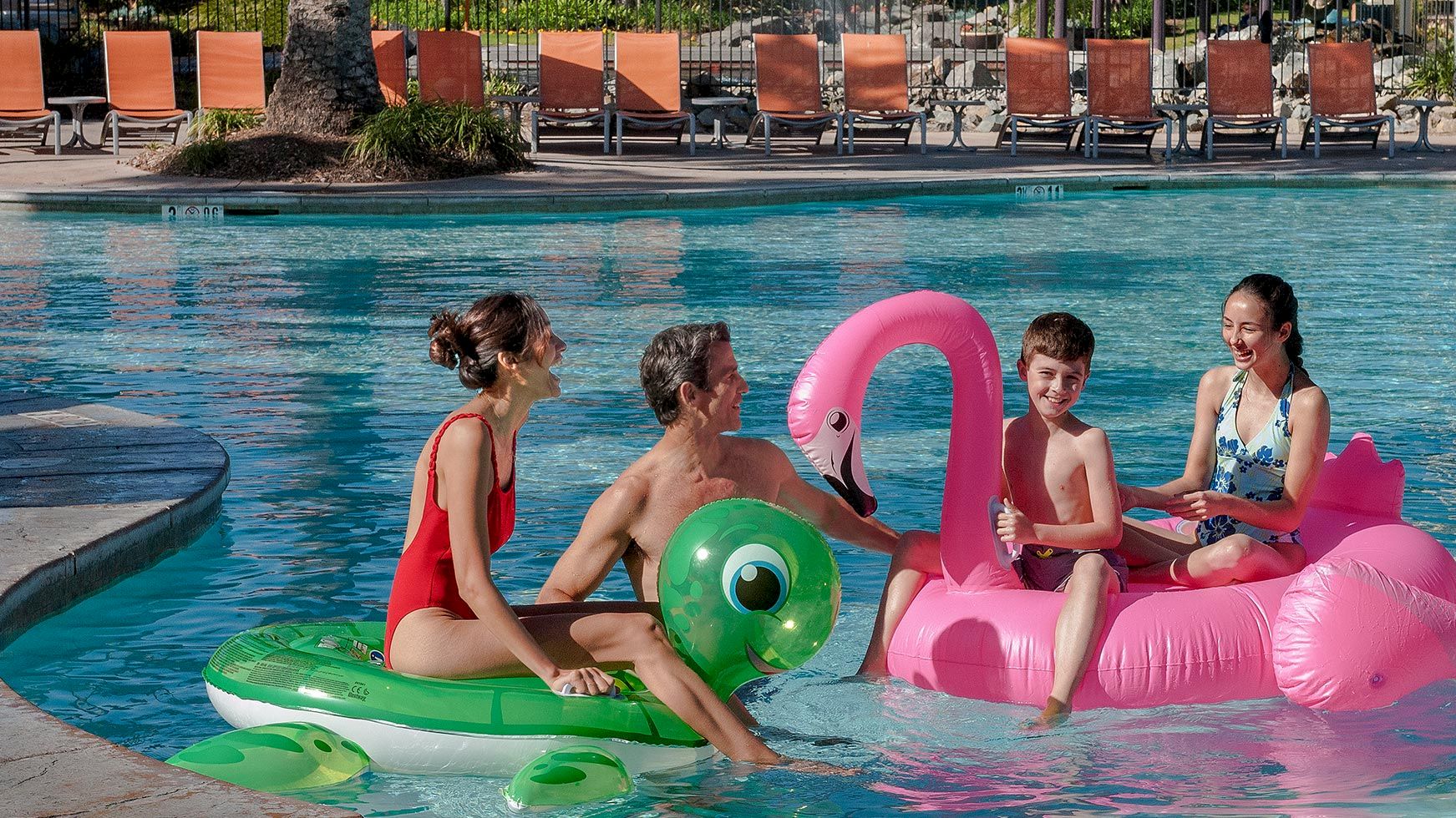 A family swims in an outdoor pool with colorful inflatable toys and a pink flamingo float on a sunny day.