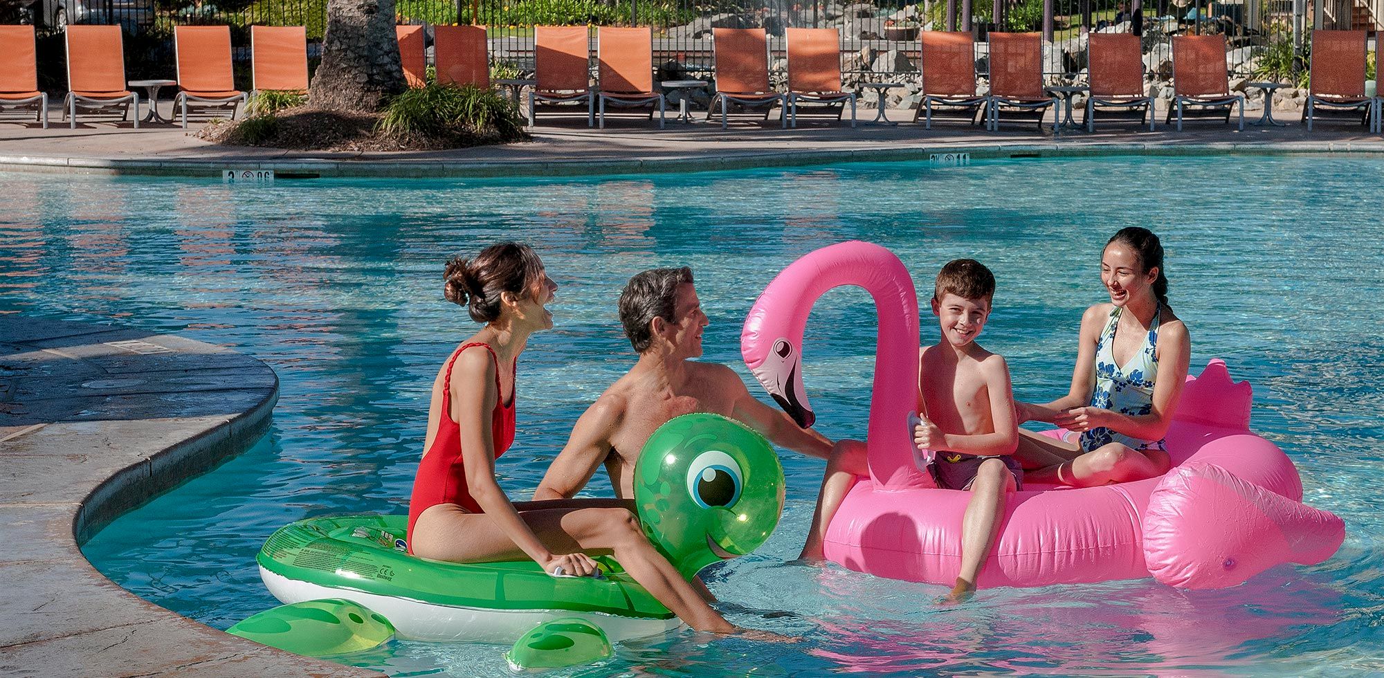 A family swims in an outdoor pool with colorful inflatable toys and a pink flamingo float on a sunny day.