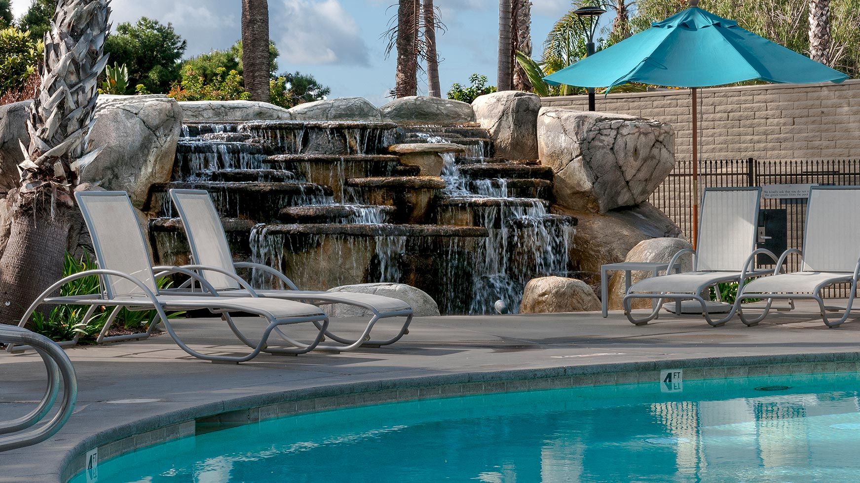 A resort pool with lounge chairs beside a multi-tier waterfall, palm trees, and a teal umbrella under a cloudy sky.