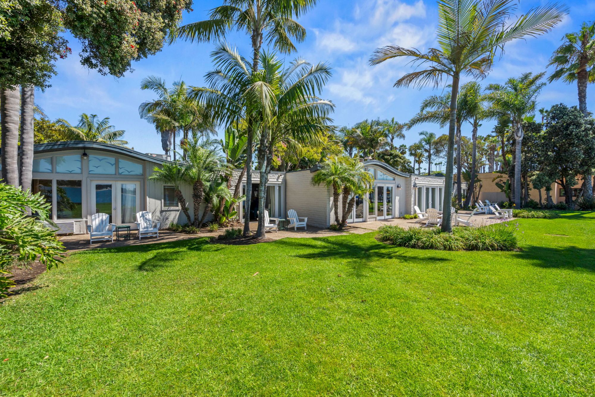 Tropical resort courtyard with palm trees, a manicured green lawn, and white cottage patios under a bright blue sky.