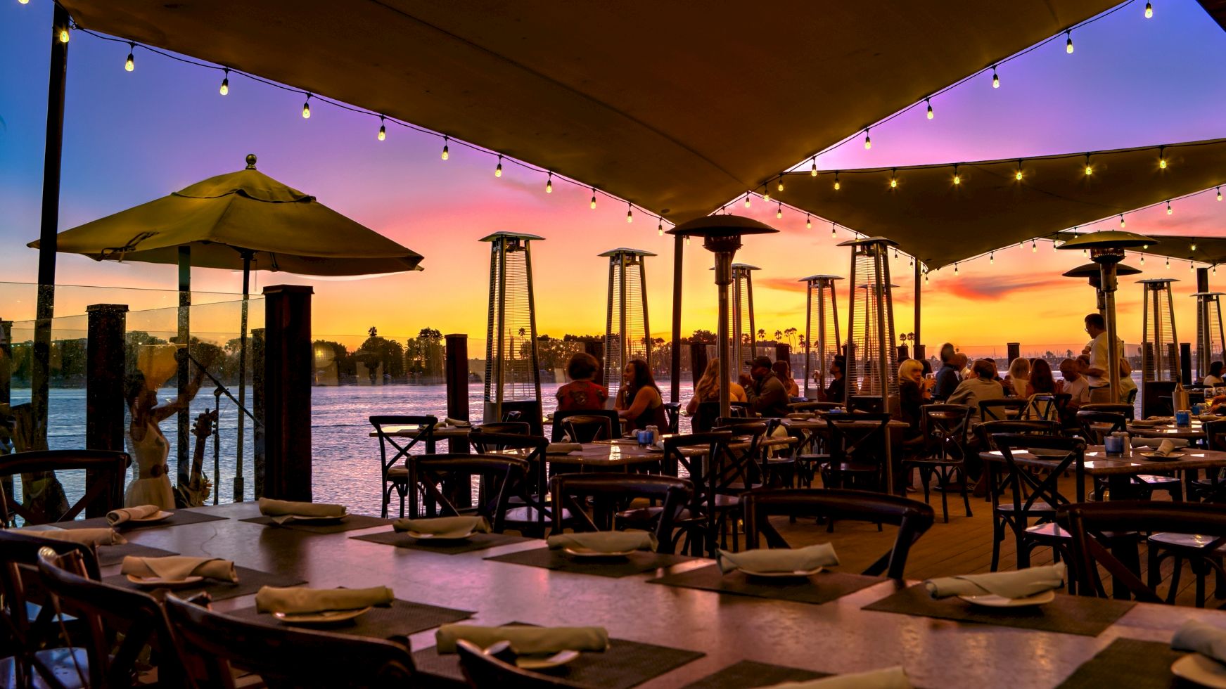 Outdoor waterfront restaurant patio at sunset with hanging string lights, umbrella shades, tabletop settings, and tall heaters.