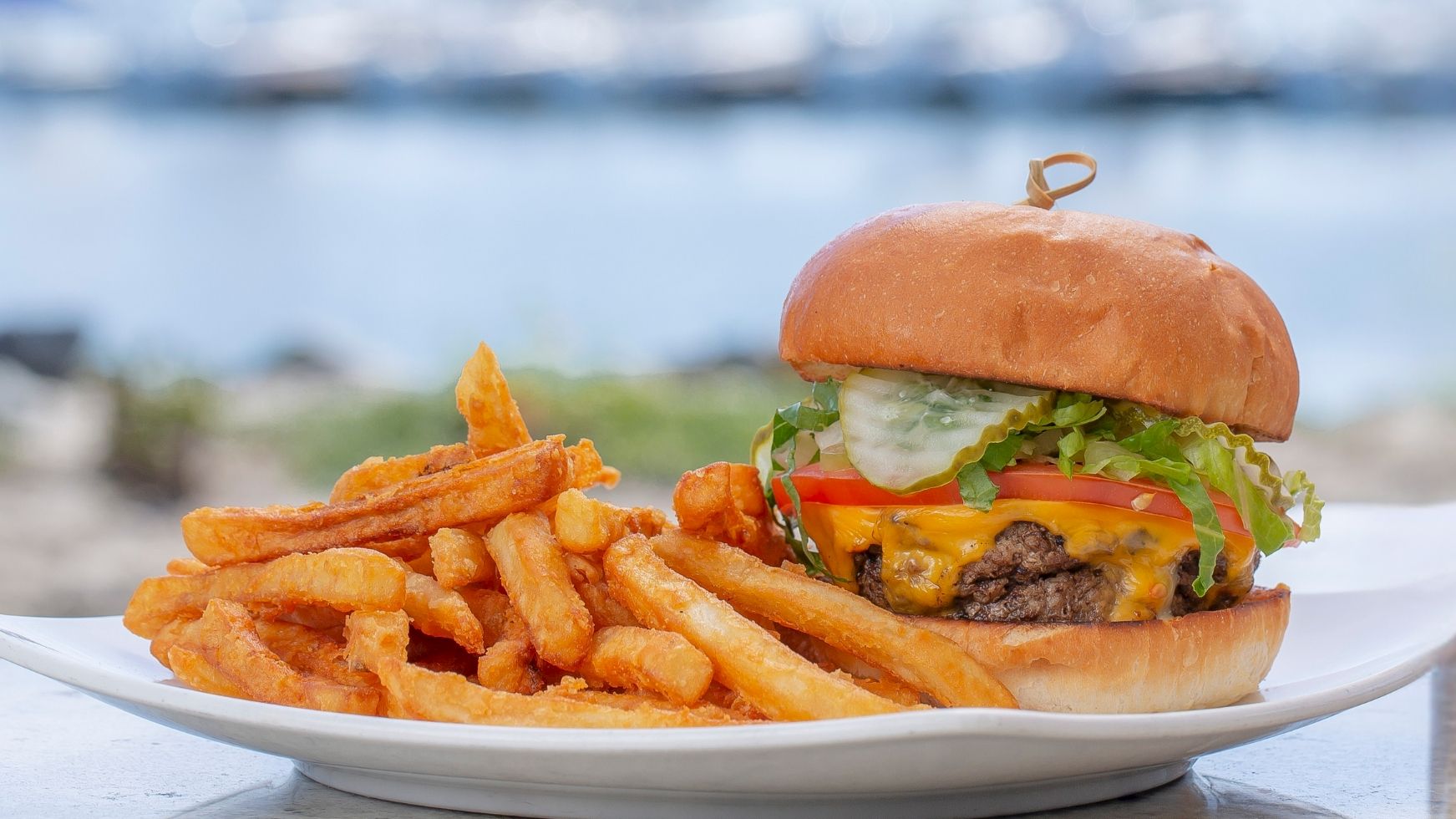 A cheeseburger with lettuce and tomato, plus thick fries on a beachside plate with the ocean blurred in the background.