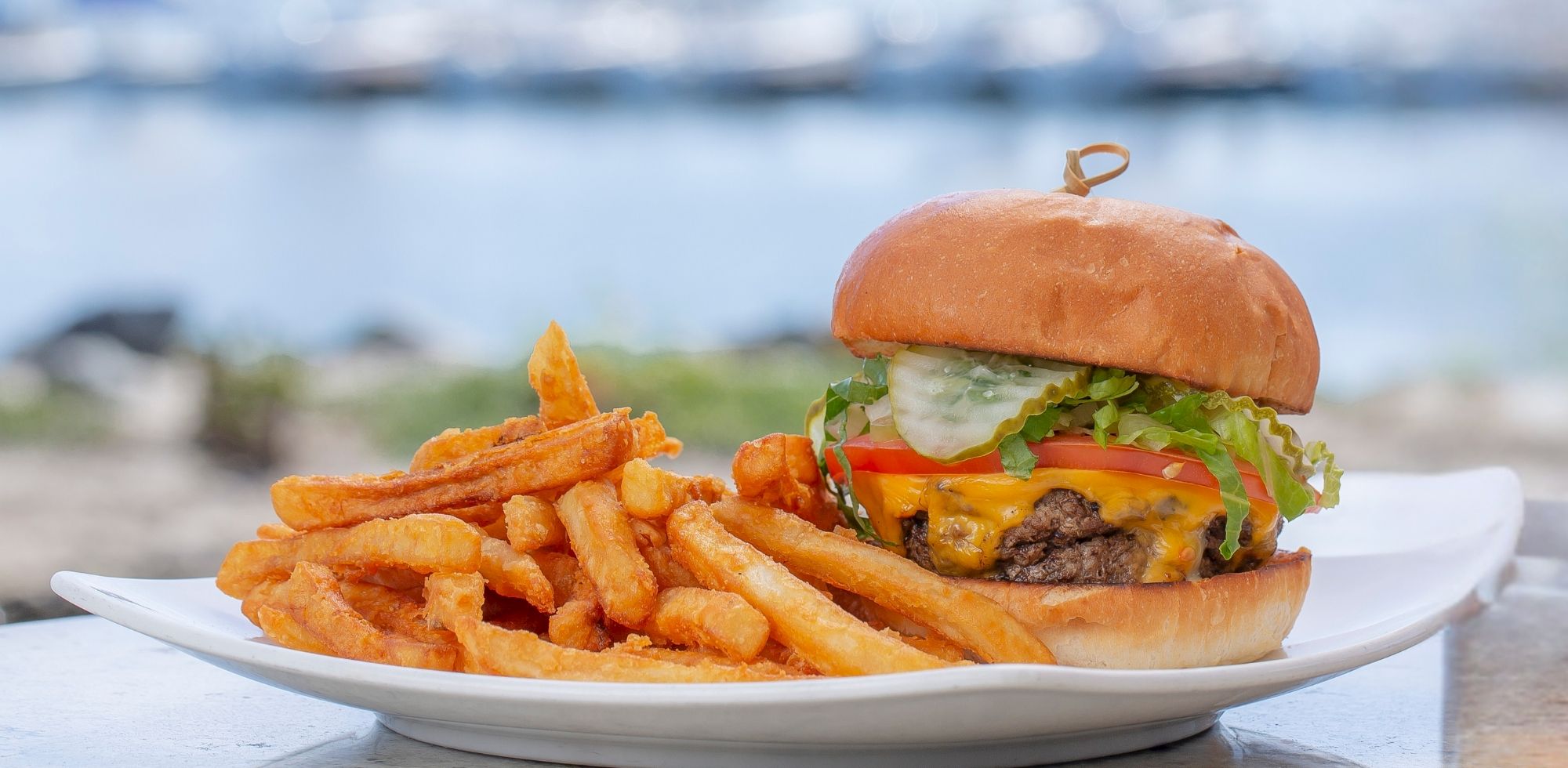 A cheeseburger with lettuce and tomato, plus thick fries on a beachside plate with the ocean blurred in the background.
