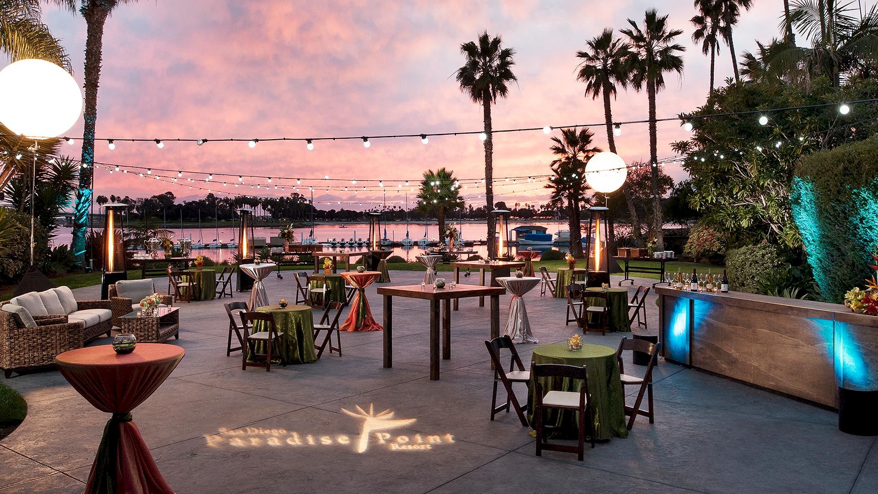 An elegant outdoor waterfront event setup at sunset with palm trees, string lights, round tables, and a lit pool nearby.
