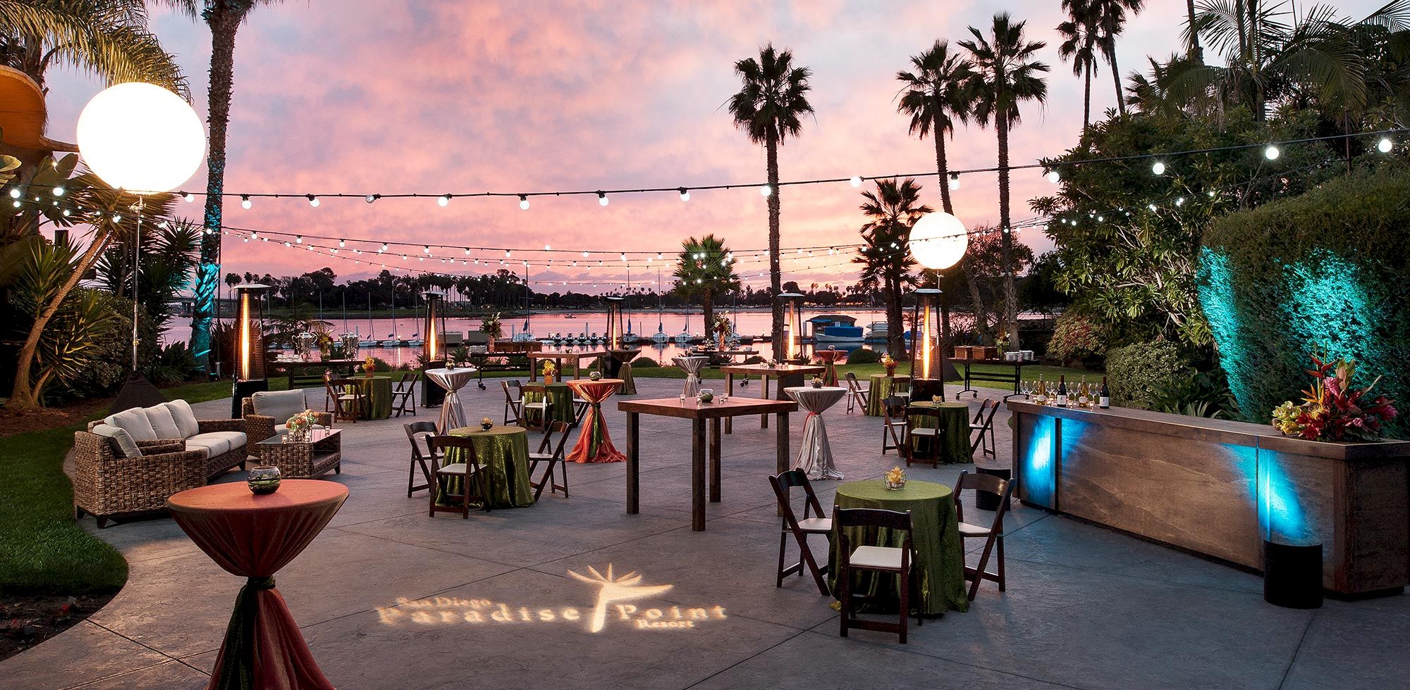 An elegant outdoor waterfront event setup at sunset with palm trees, string lights, round tables, and a lit pool nearby.