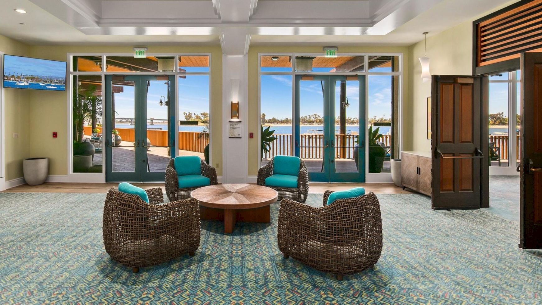 A modern lounge with wicker chairs, a round table, and large windows showing a waterfront view with palms and marina boats.