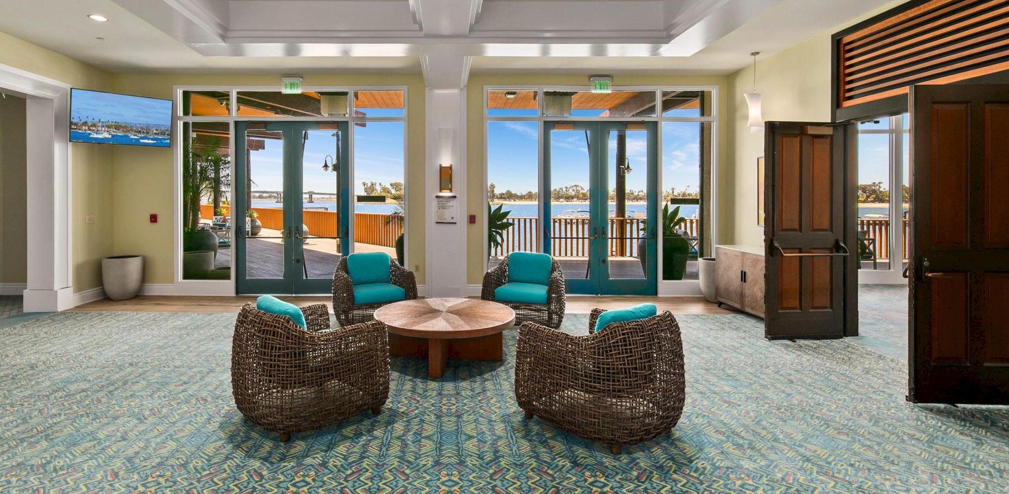 A modern lounge with wicker chairs, a round table, and large windows showing a waterfront view with palms and marina boats.