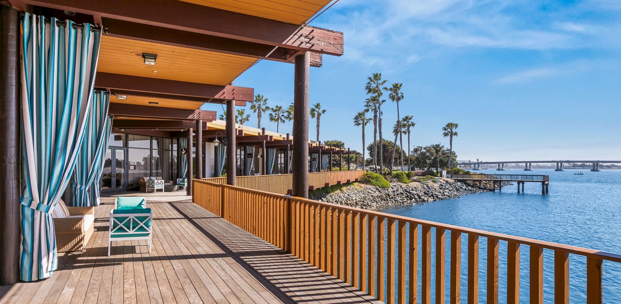 A sunny seaside patio with striped curtains, a lounge chair, wooden railings, palm trees, and blue ocean waves beyond.