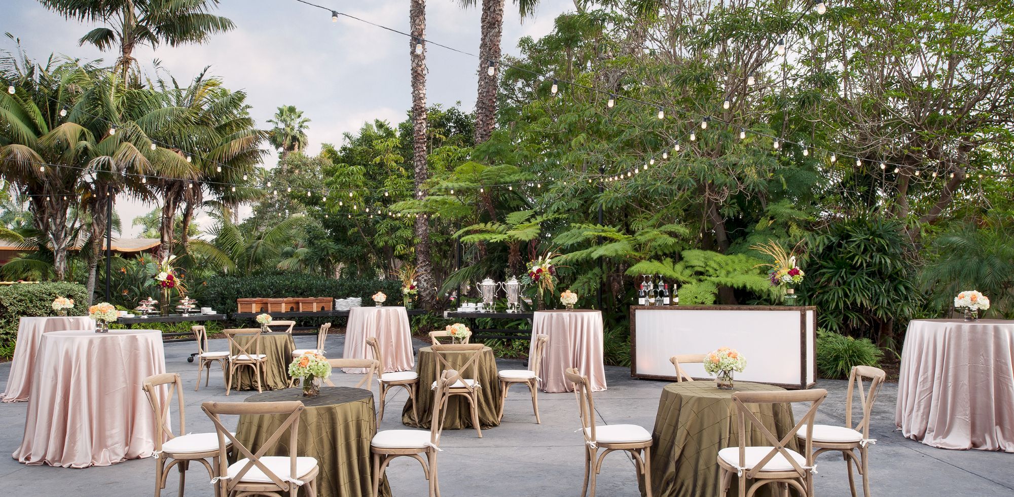 An outdoor event setup with decorated tables, chairs, floral centerpieces, string lights, and lush tropical palms and greenery.