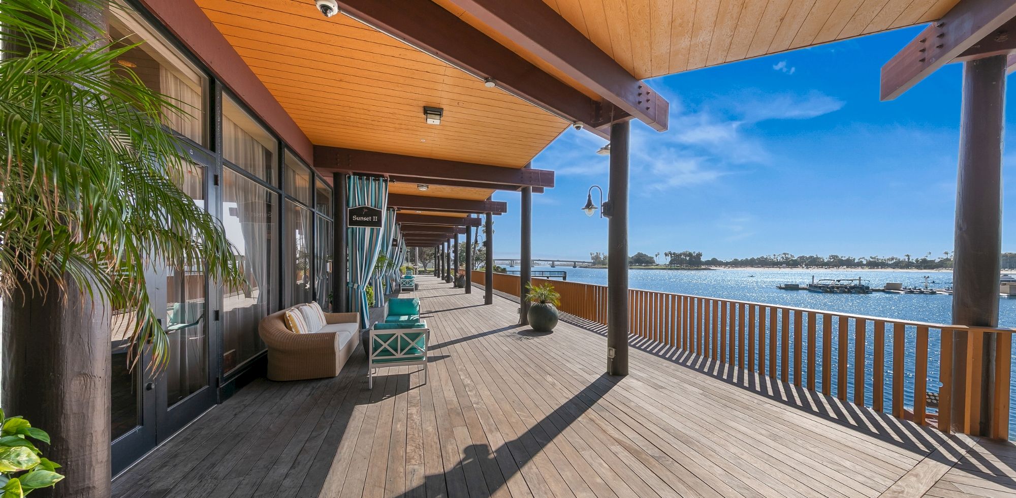 Sunny waterfront hotel deck with lounge seating, wooden pergola, and ocean view under a bright blue sky.