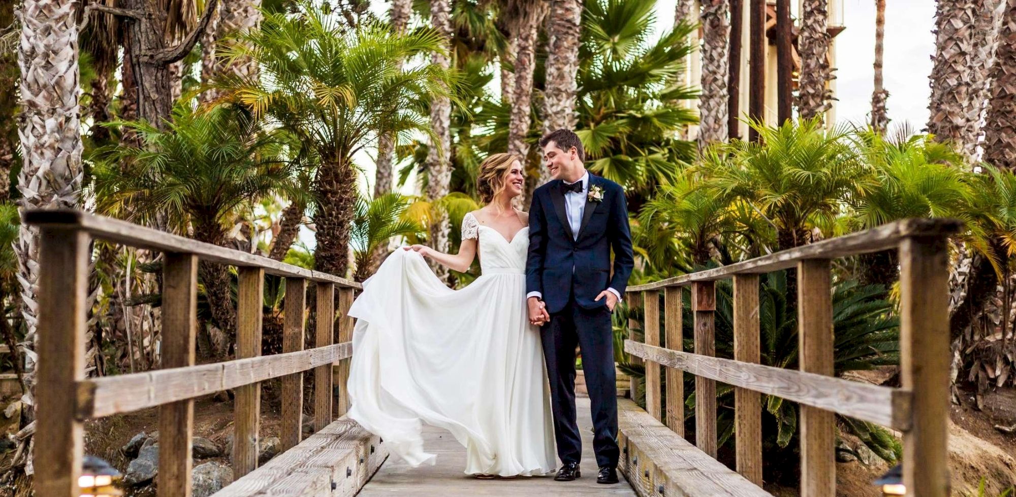 A bride in a white gown and her groom in a navy tuxedo stand on a wooden bridge amid lush palm trees, smiling at the camera.