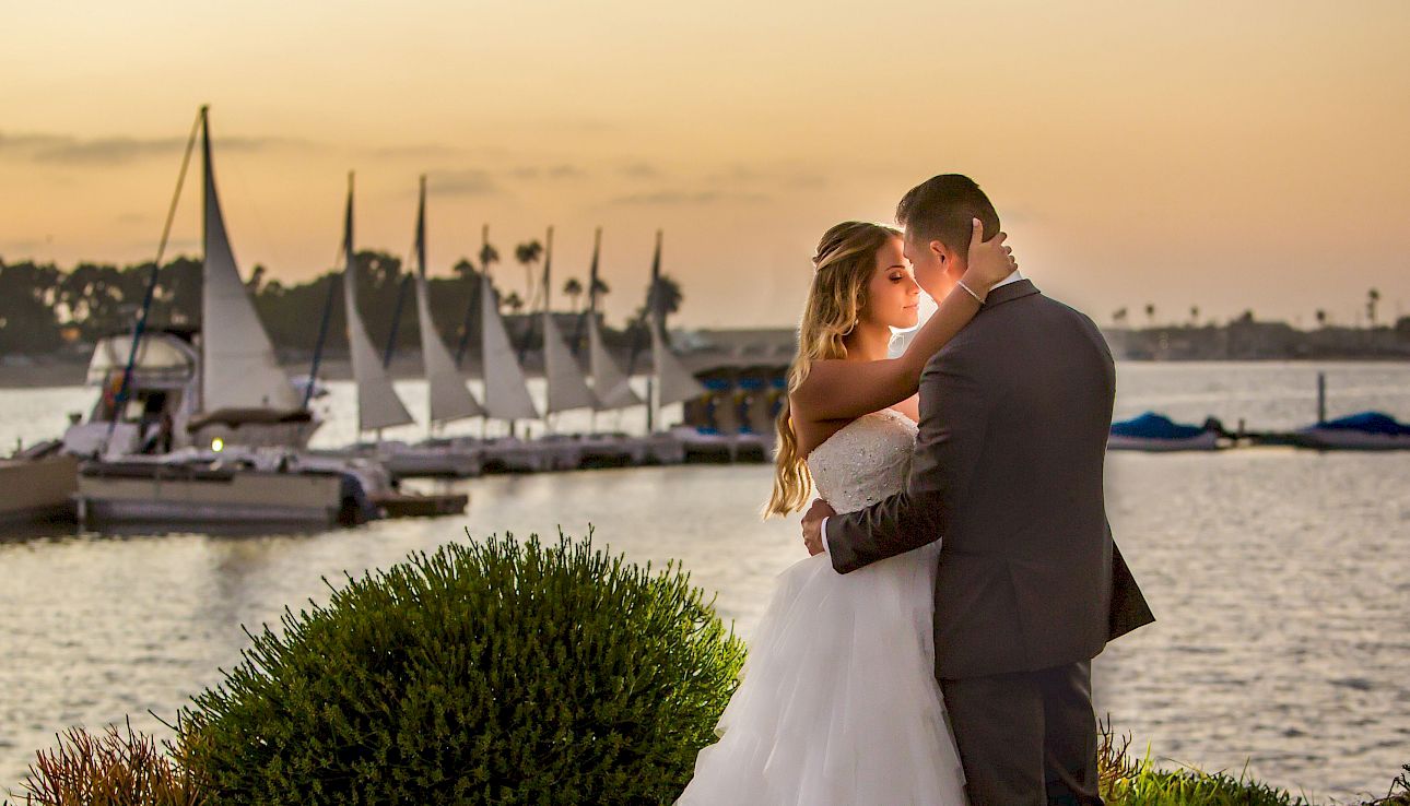 A couple in wedding attire shares a romantic kiss by the water at sunset, with sailboats moored in the background.