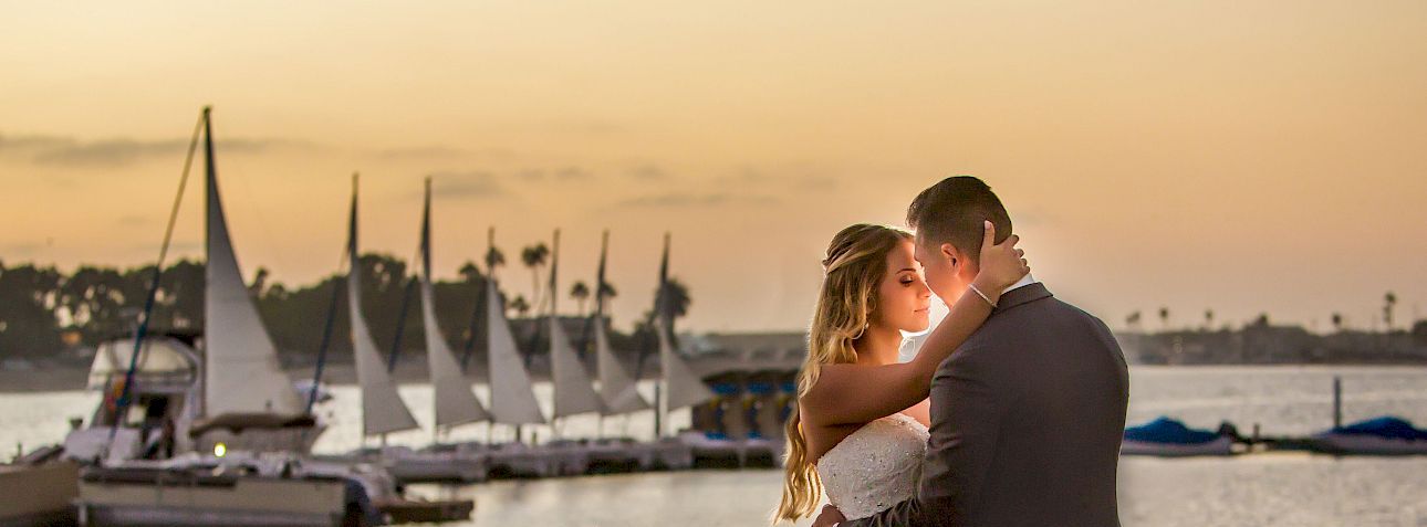 A couple in wedding attire shares a romantic kiss by the water at sunset, with sailboats moored in the background.