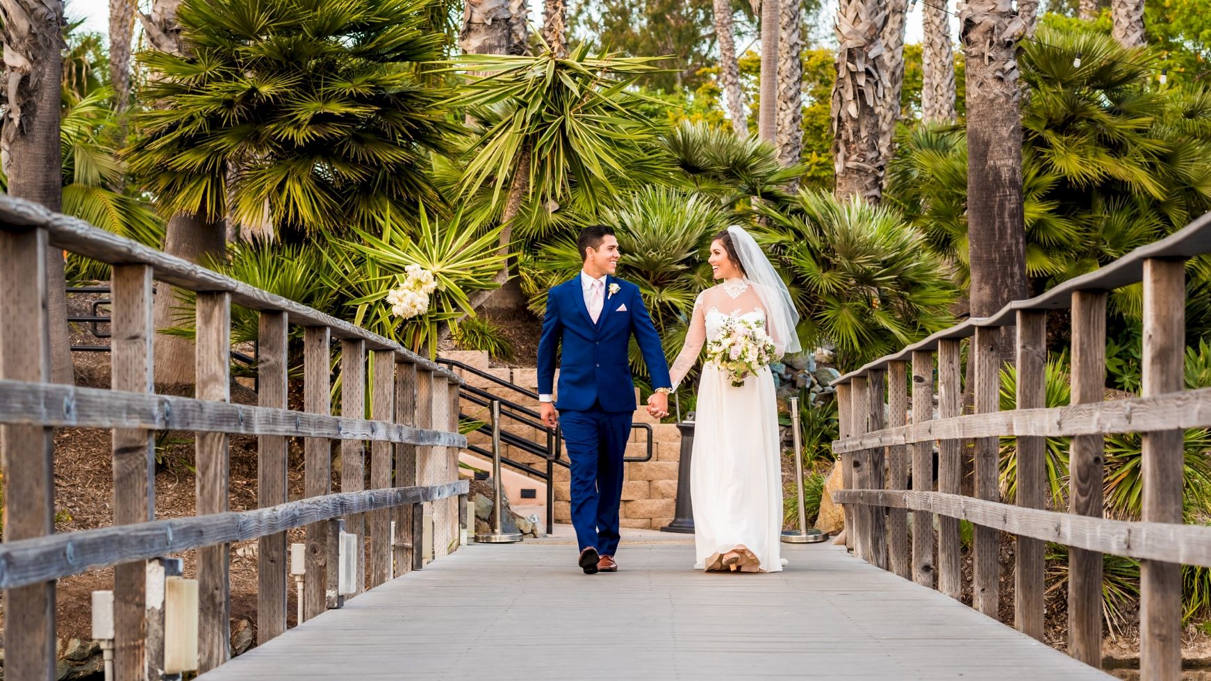 A bride and groom in blue and white walk hand-in-hand along a wooden bridge amid lush palm trees in a wedding garden.