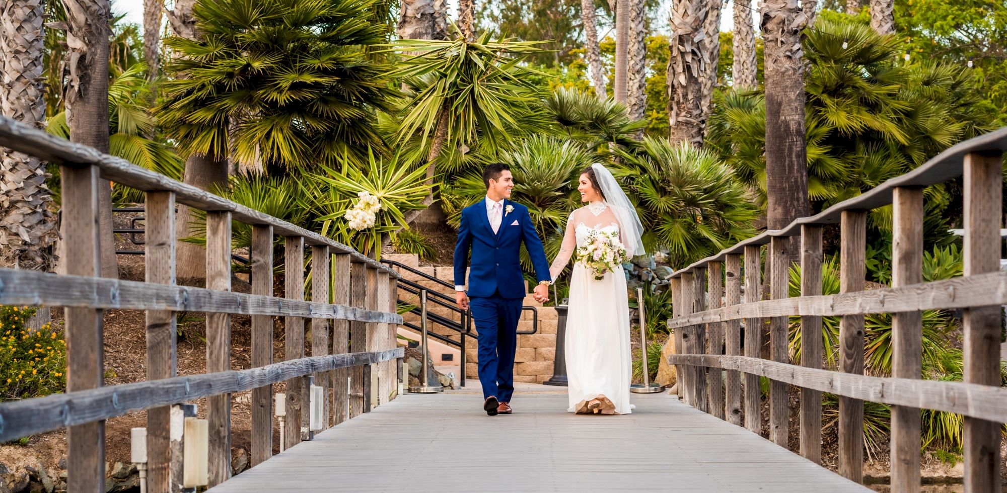A bride and groom in blue and white walk hand-in-hand along a wooden bridge amid lush palm trees in a wedding garden.