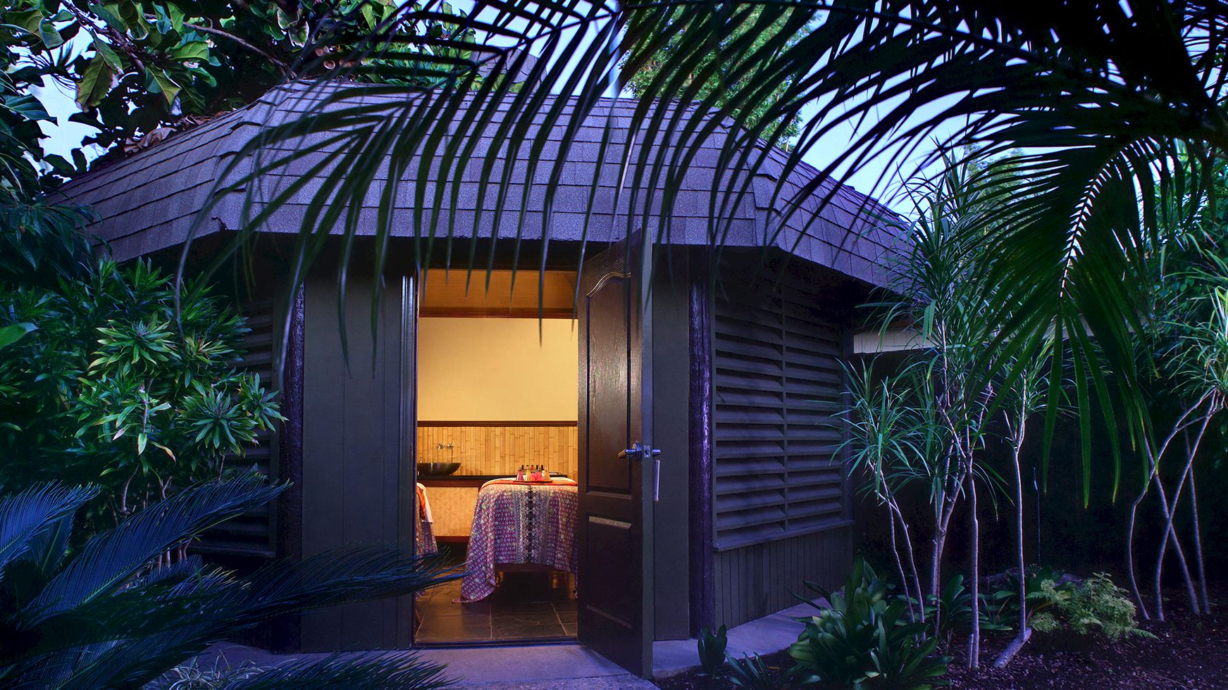 A tropical hideaway patio with lush palms and an open arched room, lit at dusk like a private dining or spa setting.
