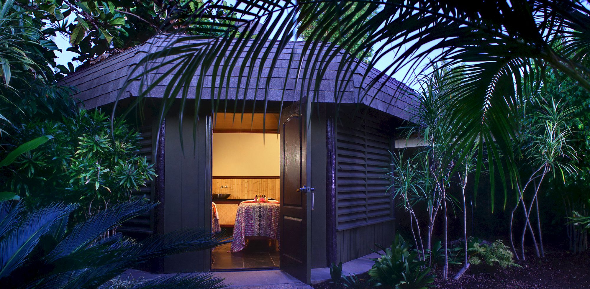 A tropical hideaway patio with lush palms and an open arched room, lit at dusk like a private dining or spa setting.