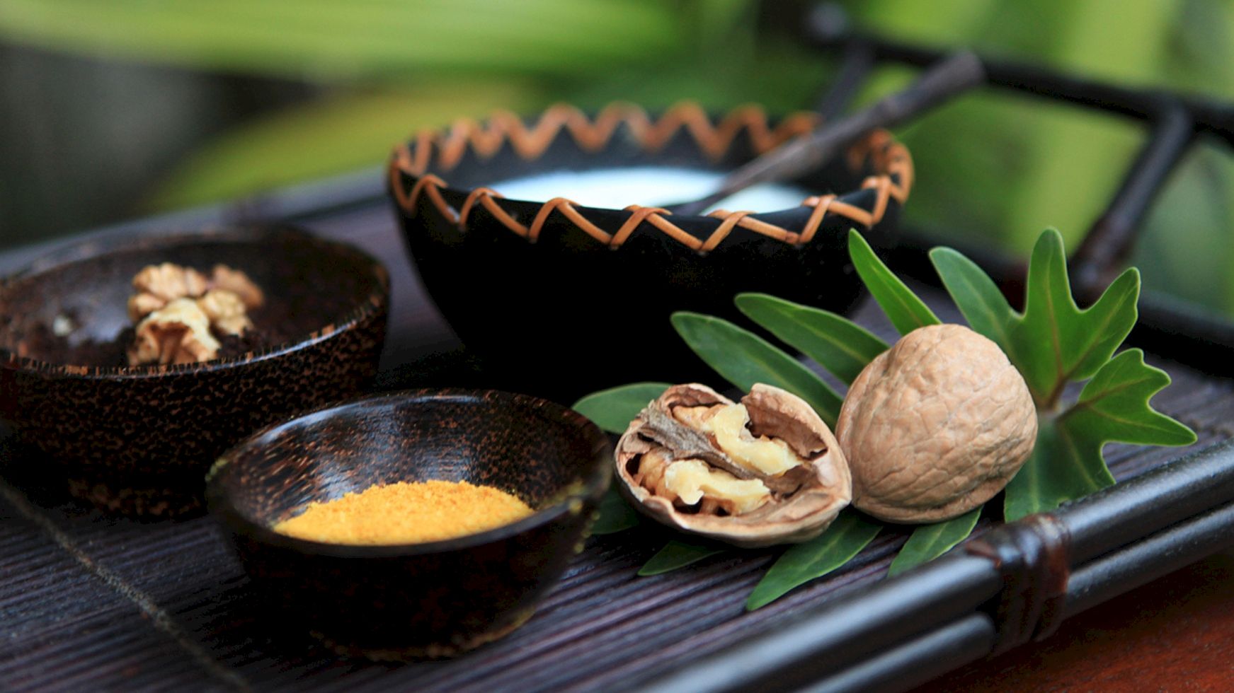A grill scene with spiced skewers, bowls of seasoning, and walnuts with fresh green leaves on a dark metal rack.