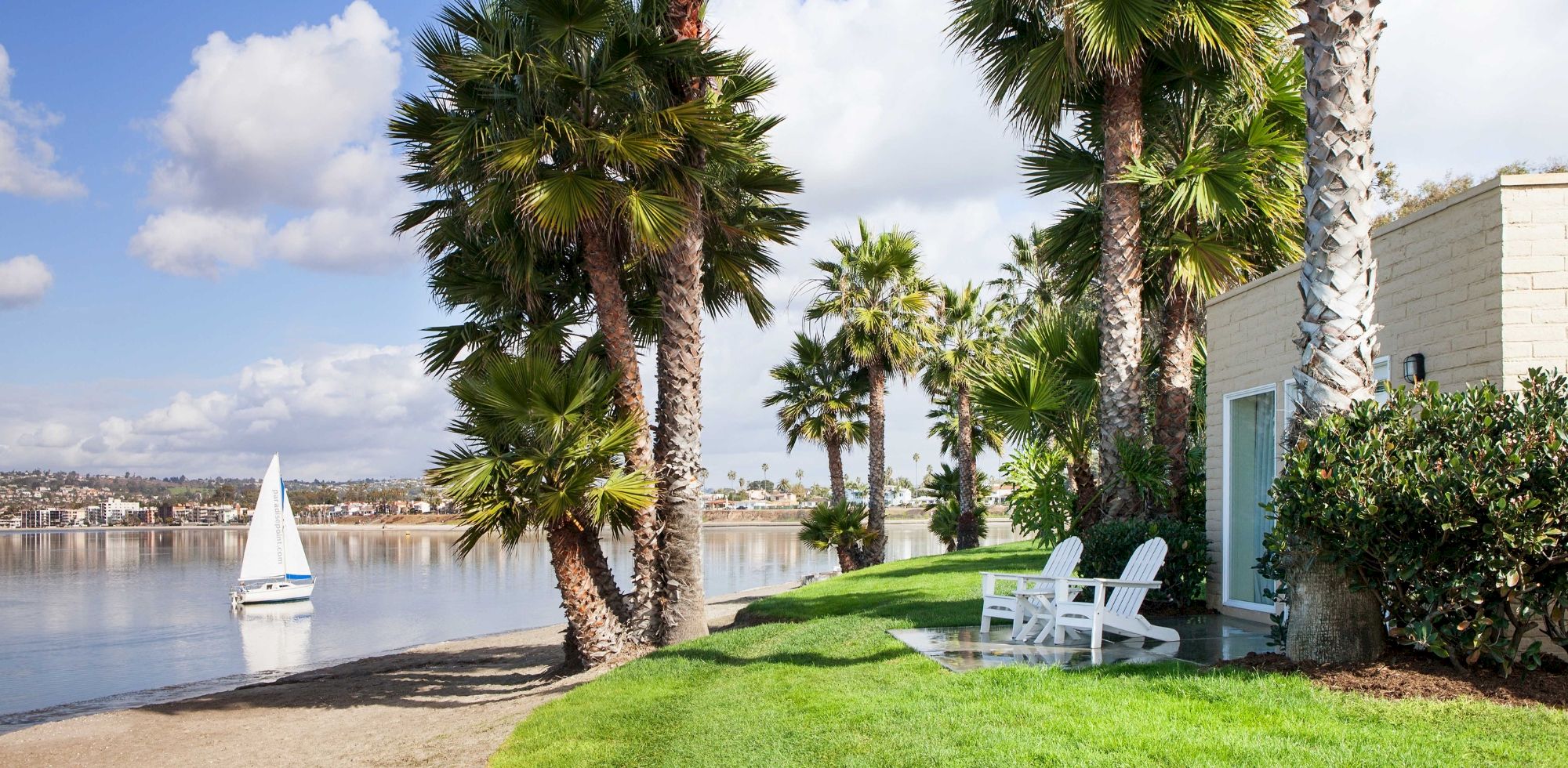 Palm trees sway by a calm lakeside, with green lawn, white patio chairs, and a sailboat on the water under blue skies.