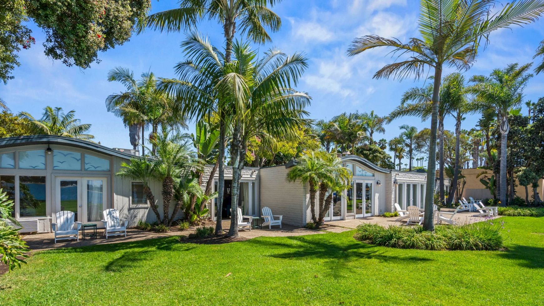 A sunny tropical resort courtyard with tall palm trees, manicured green lawn, and small guest cottages with patio chairs.