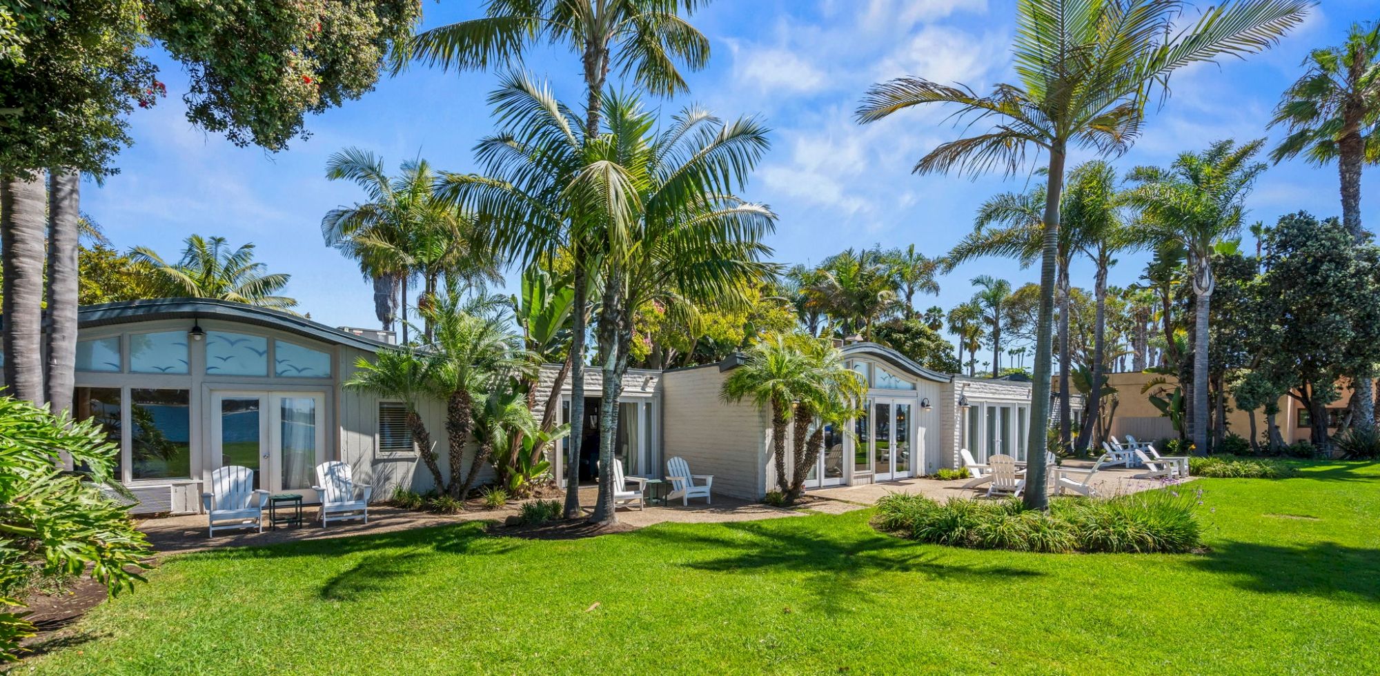 A sunny tropical resort courtyard with tall palm trees, manicured green lawn, and small guest cottages with patio chairs.