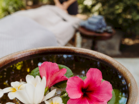 A tray/bowl of water with bright plumeria flowers sits in the foreground while a person relaxes outside in the blurred background.